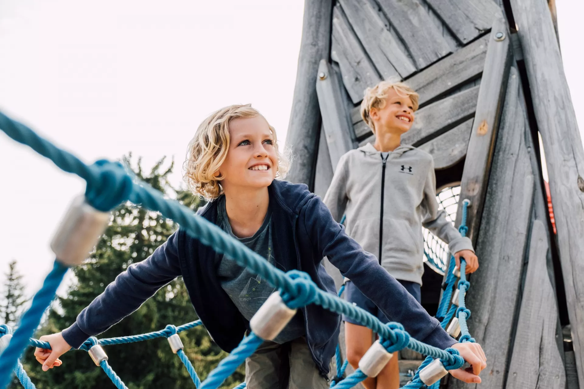 Zwei Kinder klettern lachend im Seilnetz eines hölzernen Kletterturms